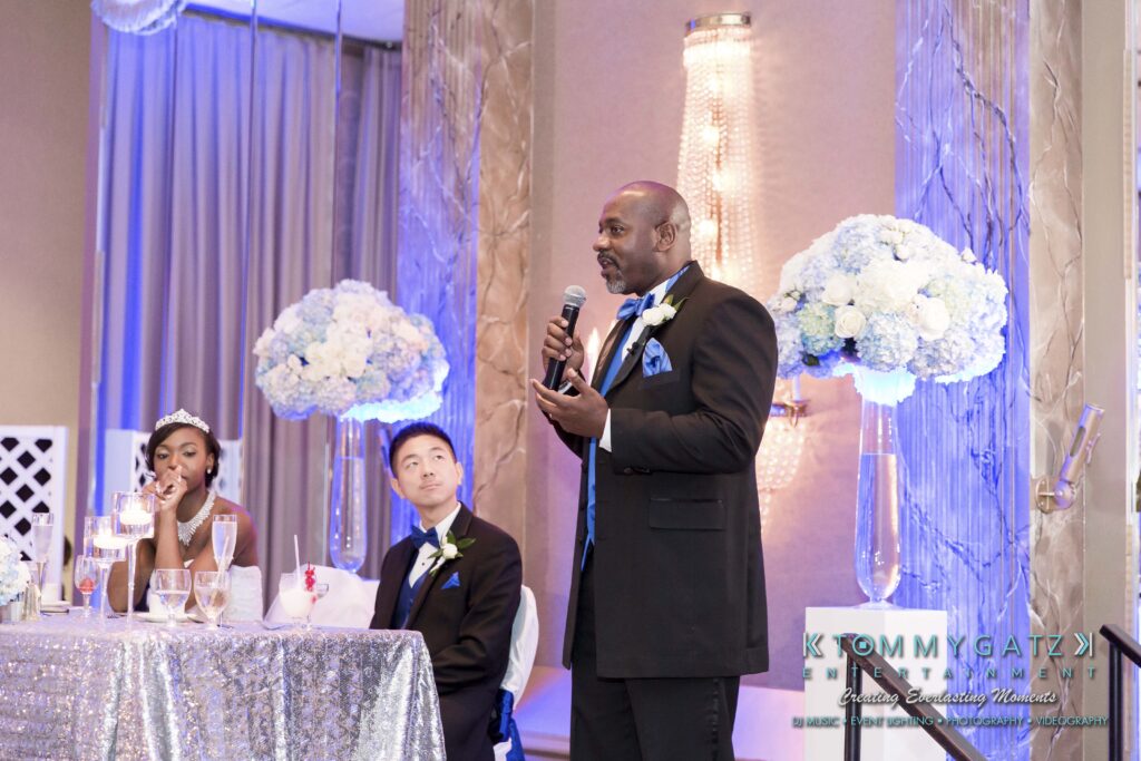 A man in a suit stands and speaks into a microphone at a wedding reception. A couple sits at a table nearby. The room is decorated with blue and white flowers and drapes.