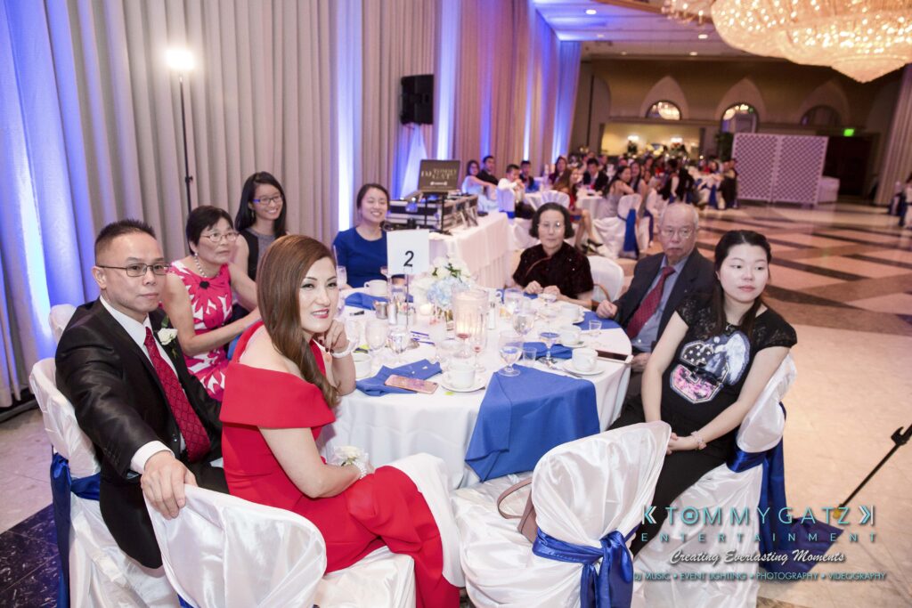 A group of people seated at round tables covered with blue and white tablecloths at an indoor event with purple lighting in the background.