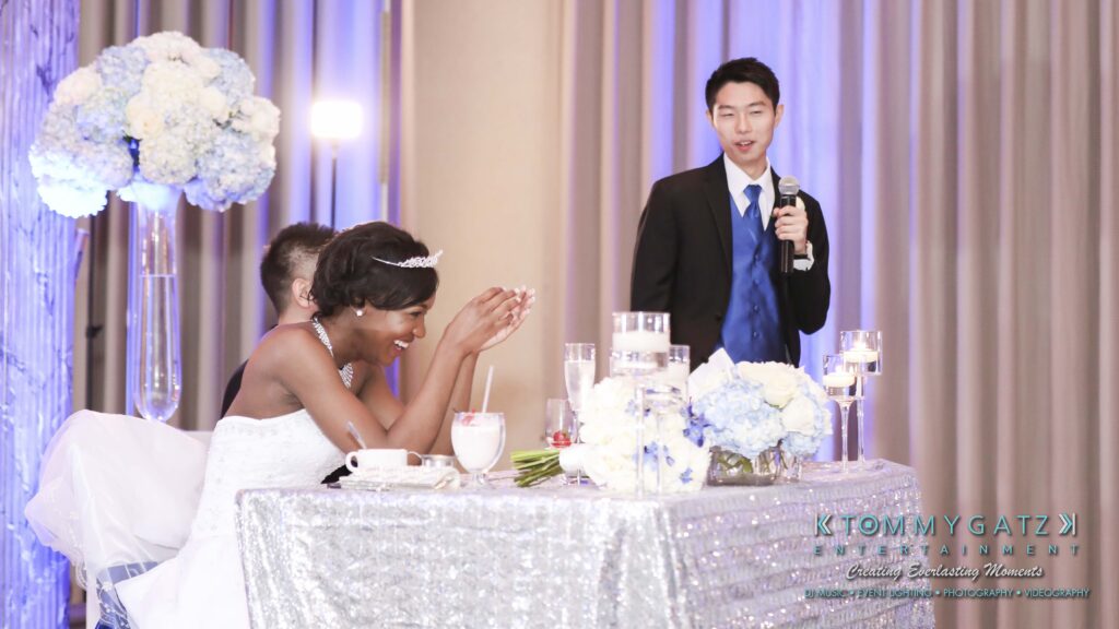 A wedding reception scene with a bride and groom seated at a table adorned with flowers and candles, while a man in a suit holds a microphone to speak to them.