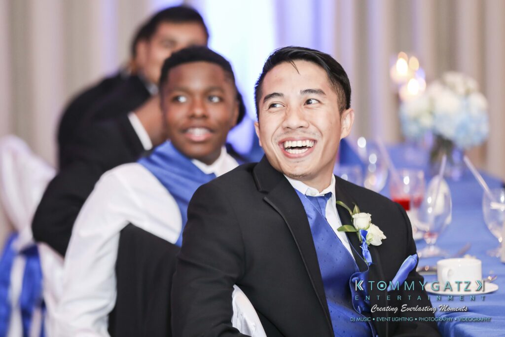 Two men in formal attire sit at a table, smiling and looking to the side. The man in the foreground wears a blue vest and boutonniere.