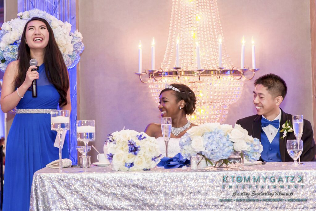 A woman in a blue dress speaks at a wedding reception, smiling. The bride and groom, seated at a decorated table, also smile. A chandelier and floral arrangement are in the background.