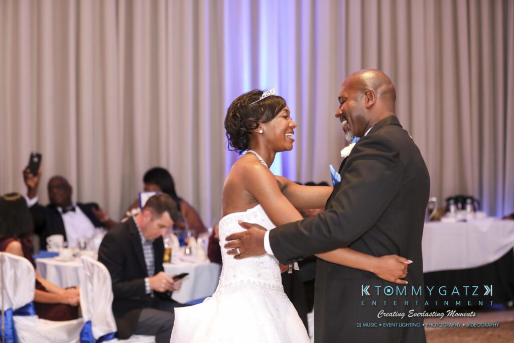 Bride and groom smiling while dancing at a wedding reception, with seated guests in the background.