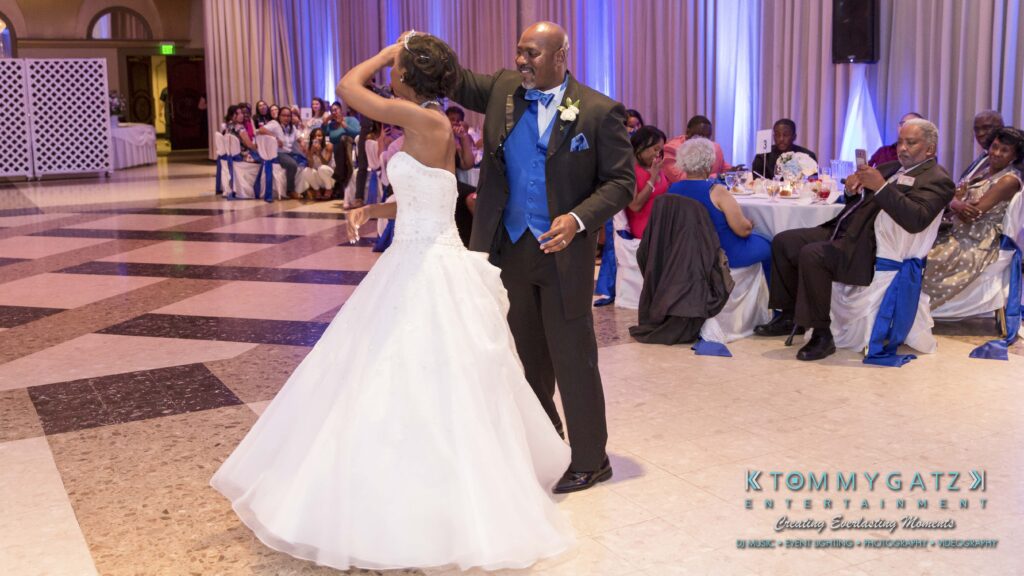 A bride and groom dance in a large hall with seated guests watching. The groom twirls the bride, who is in a white gown.