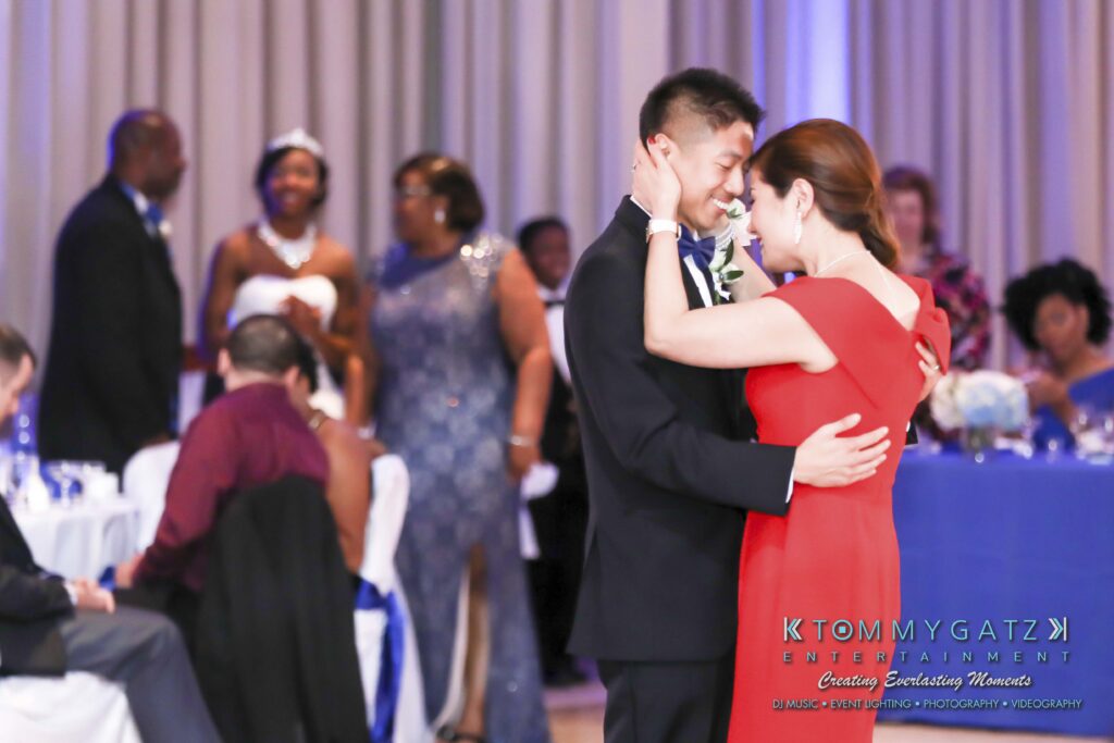 A couple dances closely in a formal setting. Other guests are seated or standing in the background, dressed in formal attire.