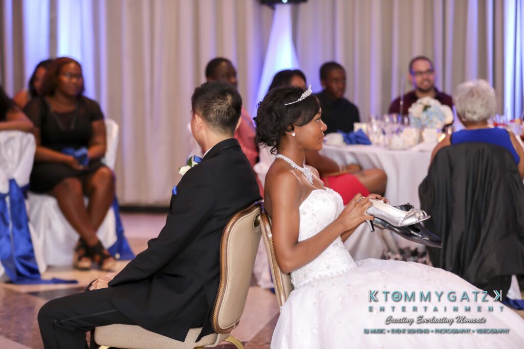 Bride and groom sitting back-to-back on chairs at a wedding reception, each holding a shoe. People are seated at tables in the background.