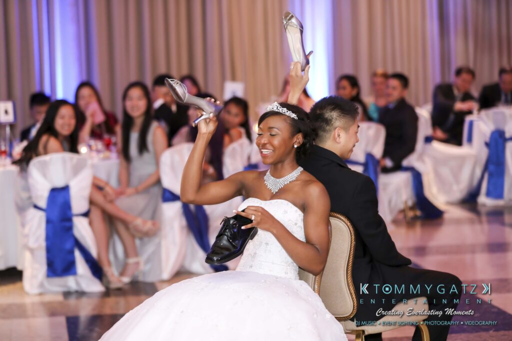Bride and groom playing the shoe game at a wedding, seated back-to-back, each holding a shoe in the air. Guests are seated in the background watching and smiling.