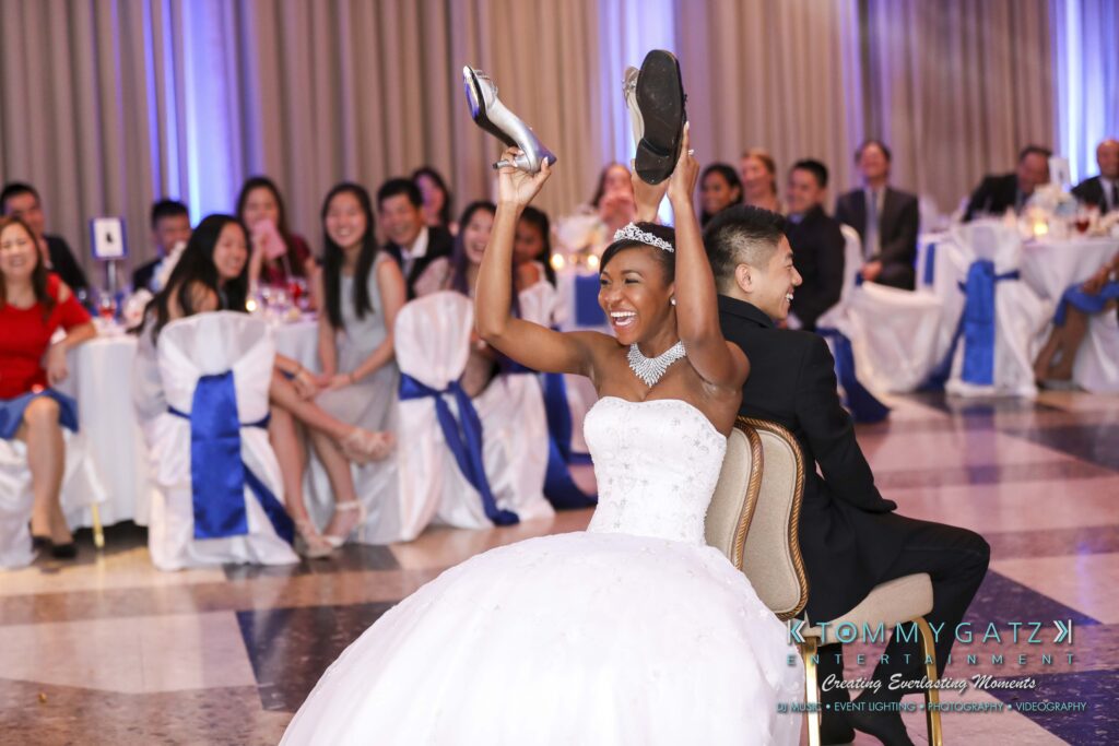 A bride and groom sit back-to-back on chairs, each holding a shoe in the air, in a room with seated guests watching.