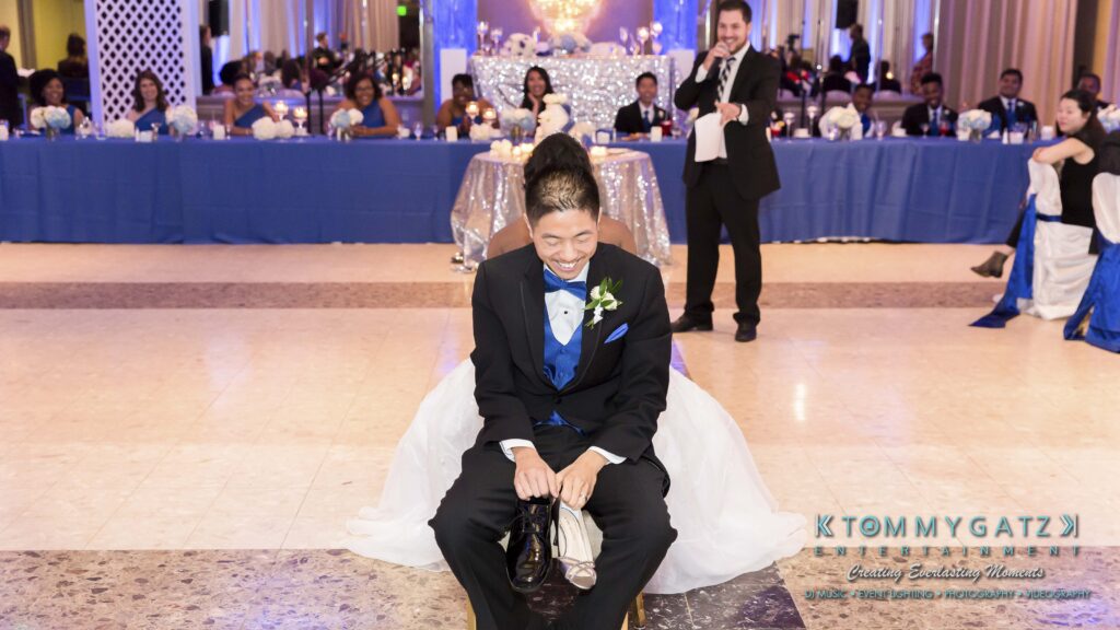 A groom in a suit and bow tie sits smiling on a dance floor with party guests in the background, while a man with a microphone stands nearby.