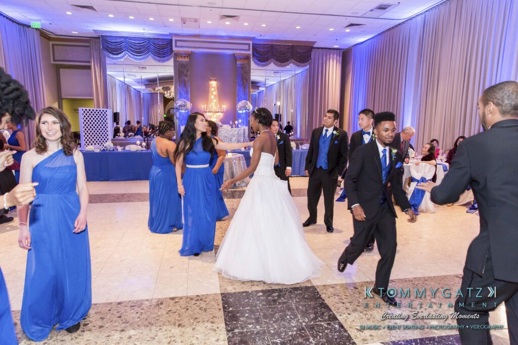 Wedding reception with people dancing. The bride in a white dress is at the center among guests in blue attire. Decor includes purple lighting and a buffet table in the background.