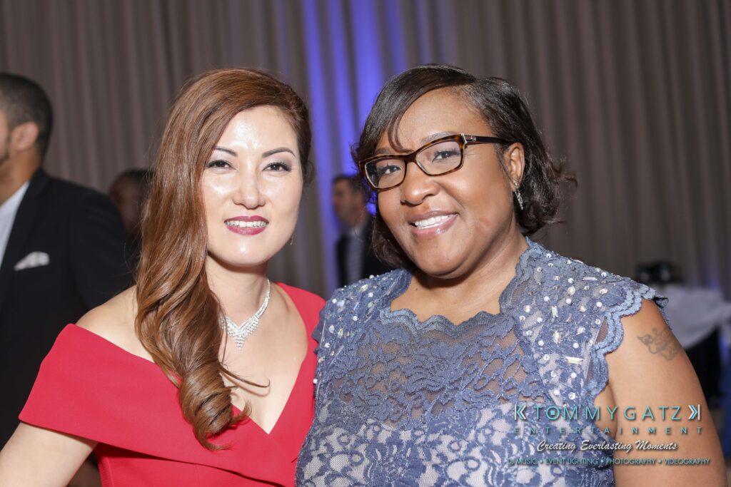 Two women smiling and posing together at an event, with one wearing a red dress and the other in a lace blue outfit.