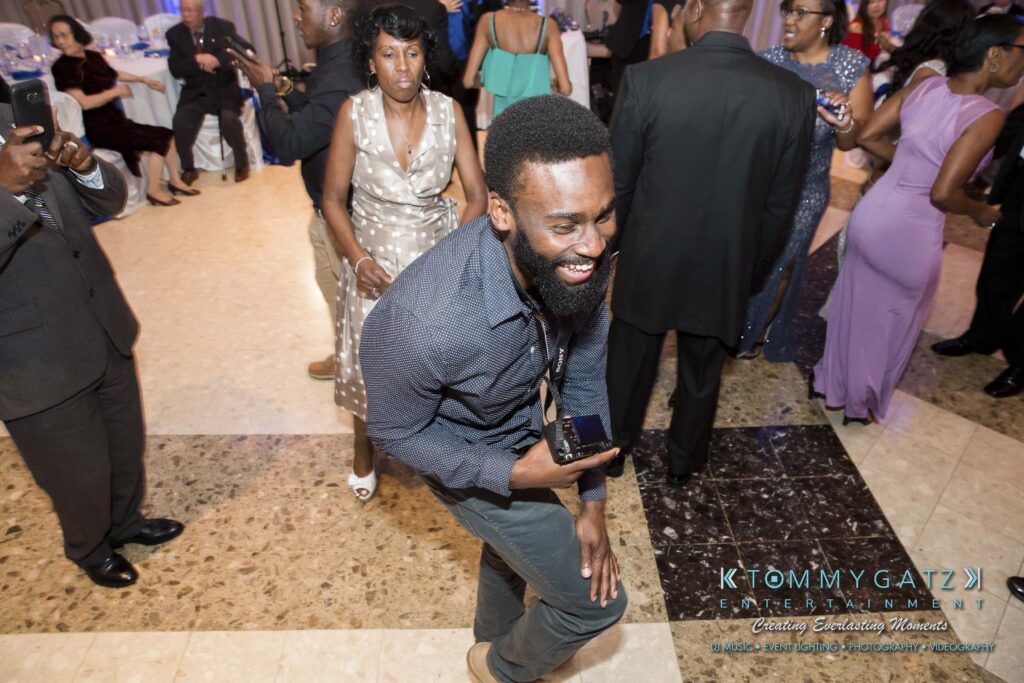 A person smiles and dances in a formal event setting. Other attendees are visible in the background, some standing and some seated.