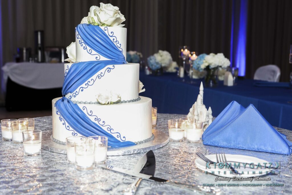 Three-tier white cake with blue drapes and floral accents, surrounded by votive candles on a silver tablecloth. A folded blue napkin and a small decorative object are nearby.