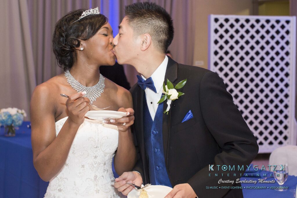 A bride and groom in formal attire share a kiss while holding small plates, with decorative elements in the background.