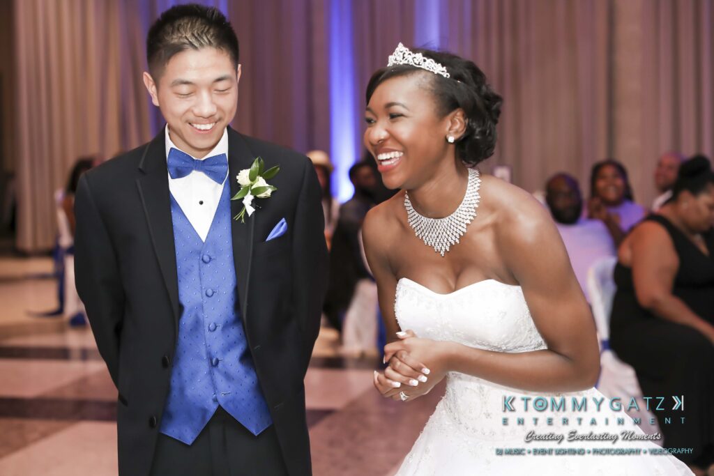 A bride and groom smiling during a wedding reception. The bride is wearing a white dress and tiara, and the groom is in a blue vest and black tuxedo. Guests are seated in the background.