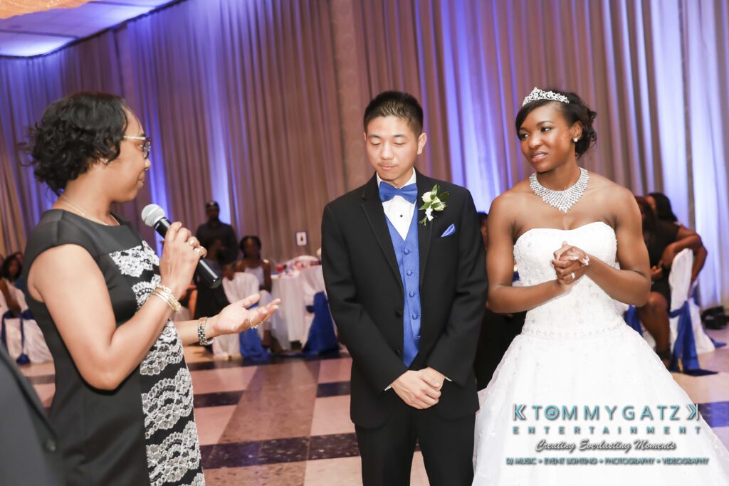 A woman speaks into a microphone at a formal event. A young man in a suit and a young woman in a white gown and tiara stand beside her, listening attentively.