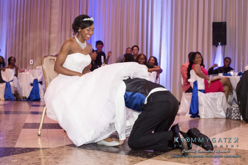 A groom searches under a bride's dress at a wedding reception. Guests watch and smile.