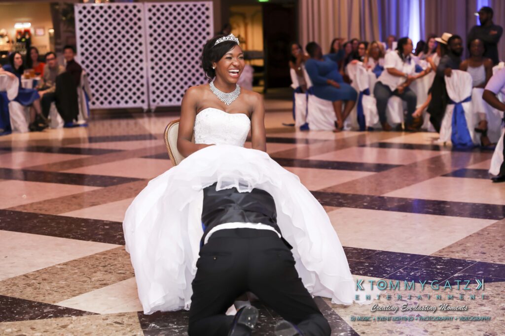 A bride smiles while seated as a groom retrieves the garter from her dress at a wedding reception. Guests are seated in the background.