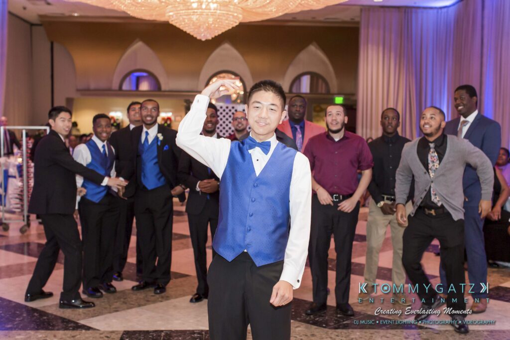 A man in a blue vest stands in front, preparing to throw a garter. A group of men behind him watches expectantly in a decorated reception hall.