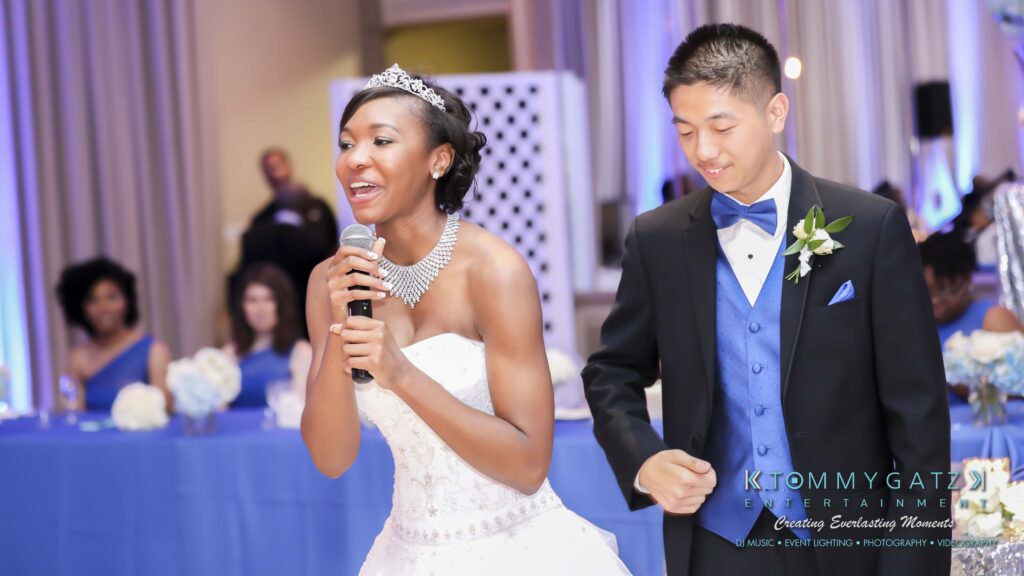 A woman in a white dress speaks into a microphone next to a man in a tuxedo. They are at an indoor event with blue-dressed guests seated in the background.