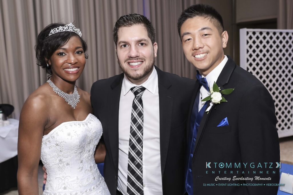 Raneka-Michael-RECEPTION-Low-Res-WM-367 - TOMMY GATZ ENTERTAINMENT Three people posing together at an event; the woman is in a white dress, and the men are in suits.