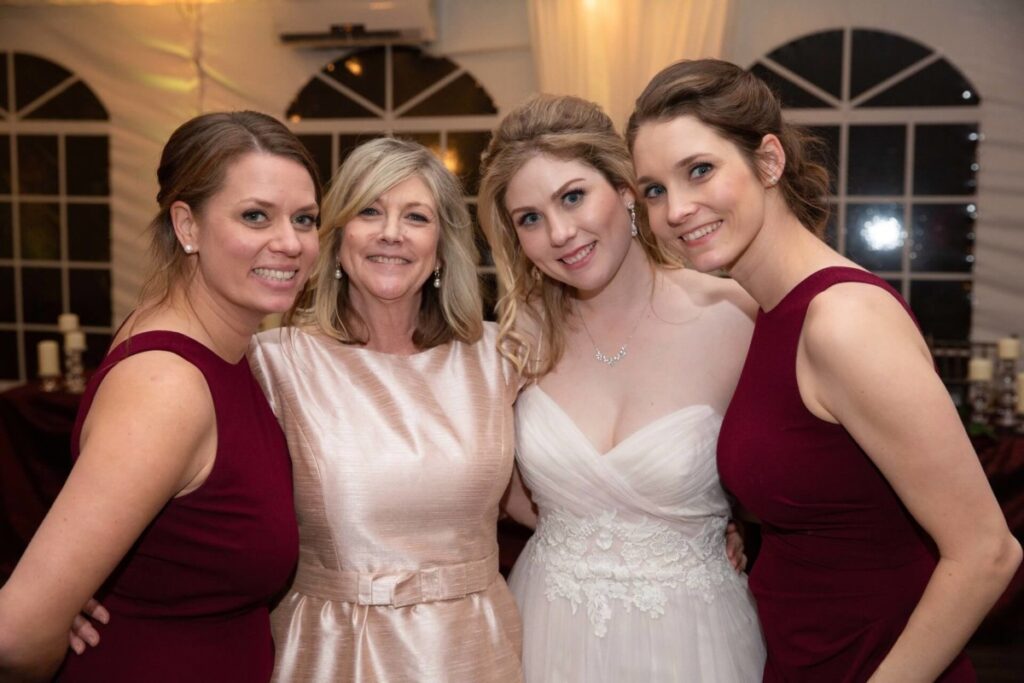 Scarlett-Michael-Wedding-Low-Res-100 - TOMMY GATZ ENTERTAINMENT Four women stand together smiling, with one in a wedding dress and the others in burgundy dresses.