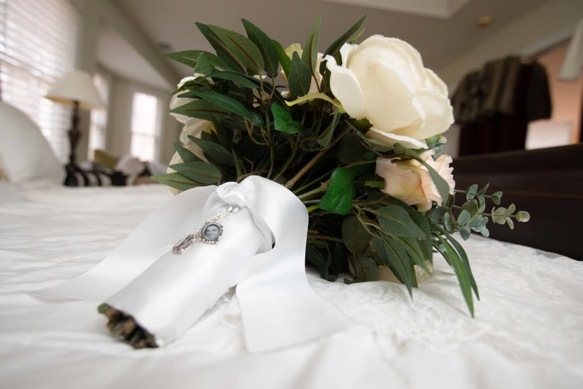 Scarlett-Michael-Wedding-Low-Res-11 - TOMMY GATZ ENTERTAINMENT A bouquet of white and pink flowers with greenery, wrapped in a white ribbon with a pendant, rests on a bed with a white bedspread.