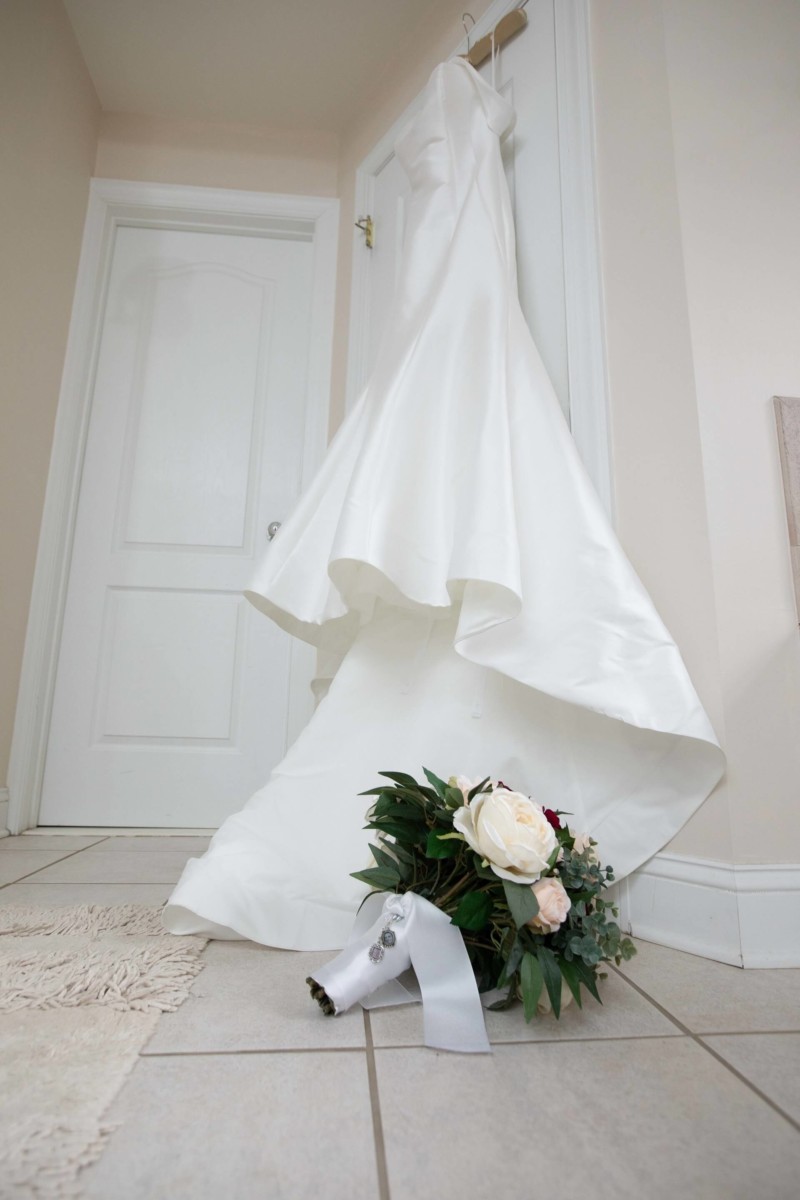 Scarlett-Michael-Wedding-Low-Res-13 - TOMMY GATZ ENTERTAINMENT White wedding dress hanging on a door, with a bridal bouquet on the floor beneath it.