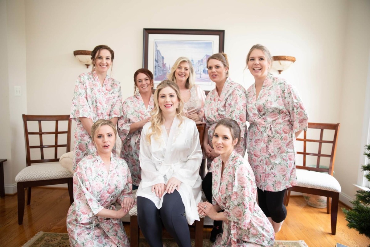 Scarlett-Michael-Wedding-Low-Res-17 - TOMMY GATZ ENTERTAINMENT A bride in a white robe sits with seven bridesmaids in matching floral robes, posing indoors in front of a painting.