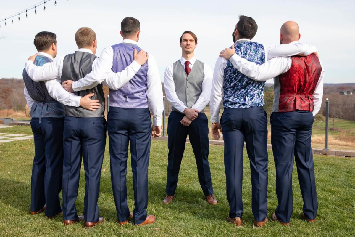 Scarlett-Michael-Wedding-Low-Res-2 - TOMMY GATZ ENTERTAINMENT A group of six men in dress shirts and colorful waistcoats stands on grass; five have arms around each other, facing away, while one faces them.