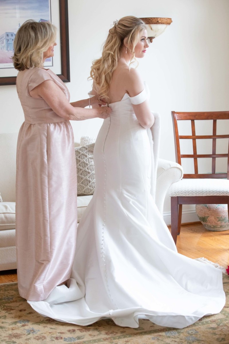 Scarlett-Michael-Wedding-Low-Res-21 - TOMMY GATZ ENTERTAINMENT A woman helps a bride adjust her white wedding dress in a room with a couch and chairs.