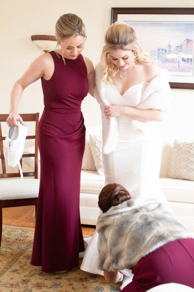 Scarlett-Michael-Wedding-Low-Res-23 - TOMMY GATZ ENTERTAINMENT Two women assist a bride in a white gown with her train. One holds white shoes, and another crouches to adjust the dress. A painting hangs on the cream-colored wall behind them.