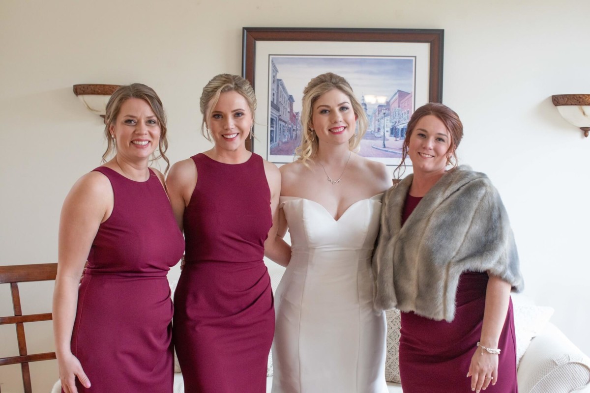 Scarlett-Michael-Wedding-Low-Res-25 - TOMMY GATZ ENTERTAINMENT Four women posing indoors, with one in a white dress and the others in burgundy dresses. A painting hangs on the wall behind them.