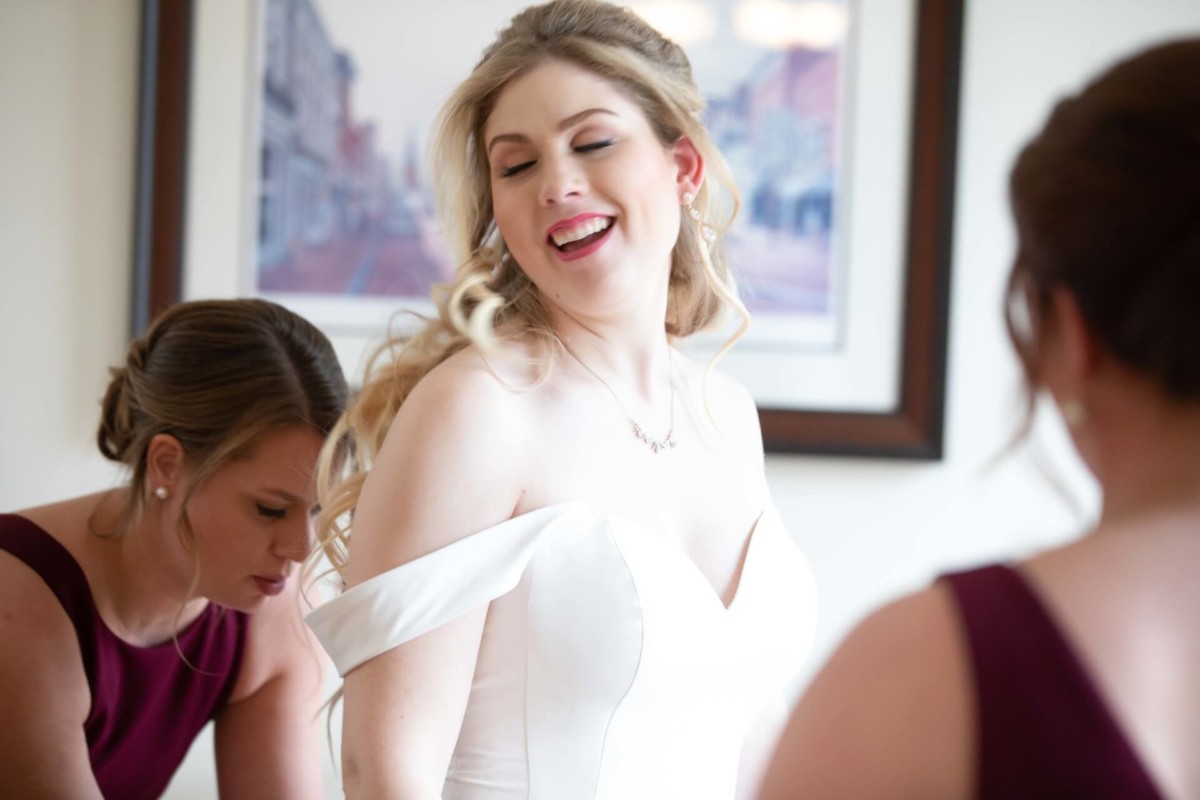 Scarlett-Michael-Wedding-Low-Res-29 - TOMMY GATZ ENTERTAINMENT Bride smiling with eyes closed, wearing an off-shoulder white dress; two bridesmaids in burgundy dresses adjust her gown.