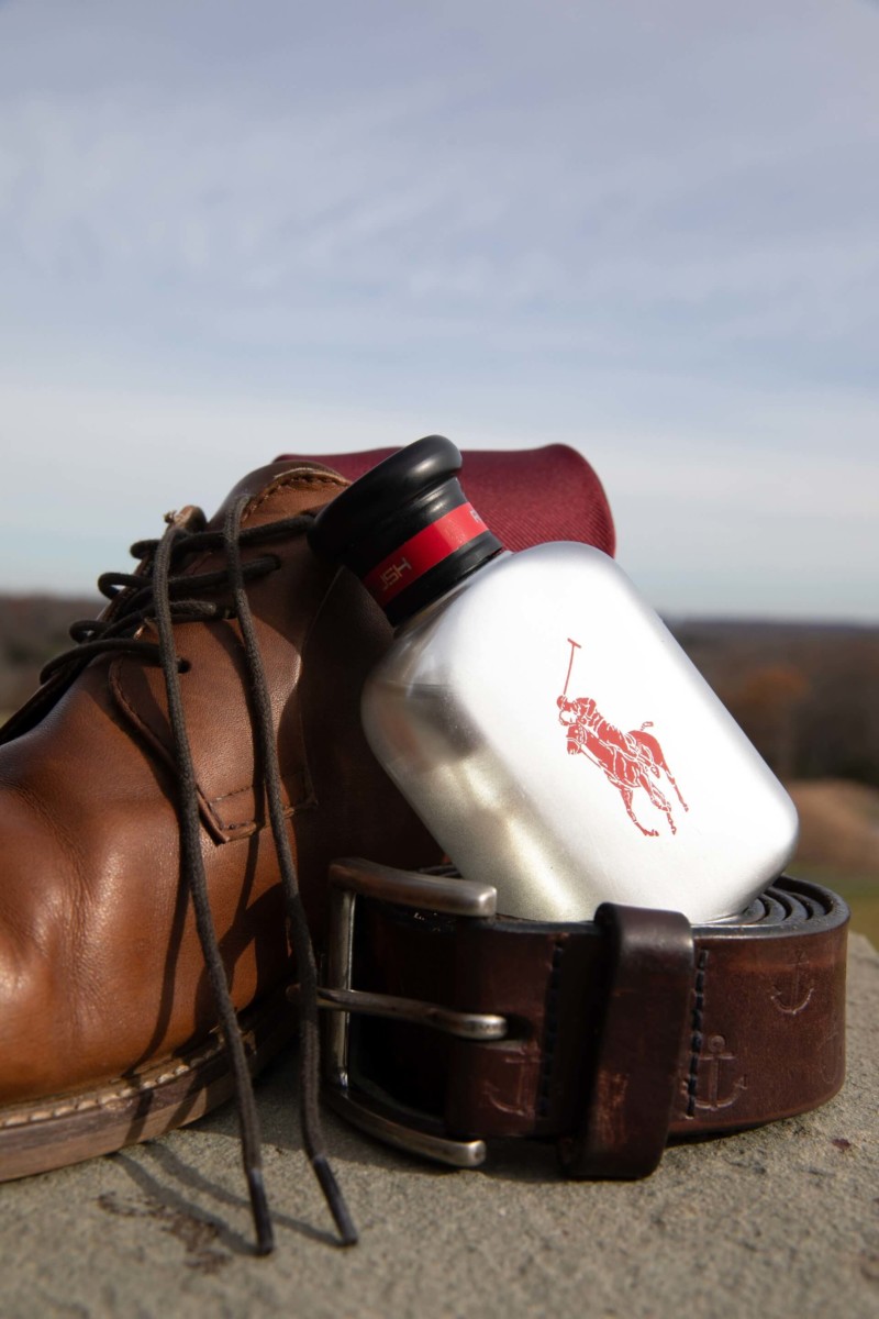 Scarlett-Michael-Wedding-Low-Res-3 - TOMMY GATZ ENTERTAINMENT Brown leather boot and belt with a silver flask featuring a red logo, set against an outdoor background.
