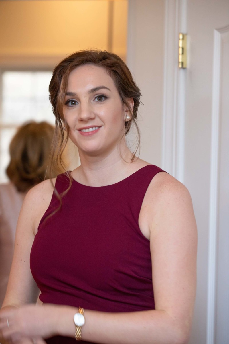 Scarlett-Michael-Wedding-Low-Res-30 - TOMMY GATZ ENTERTAINMENT Woman in a burgundy dress standing in a room, smiling slightly, with a watch on her wrist.