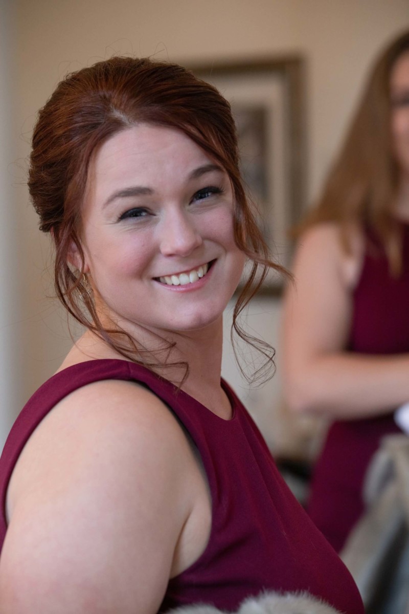 Scarlett-Michael-Wedding-Low-Res-31 - TOMMY GATZ ENTERTAINMENT Woman with auburn hair in a burgundy dress smiles at the camera, with another person slightly blurred in the background.