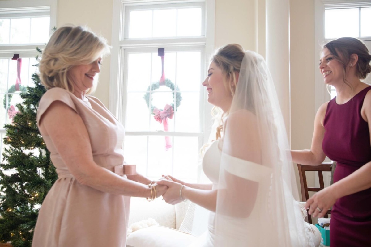 Scarlett-Michael-Wedding-Low-Res-34 - TOMMY GATZ ENTERTAINMENT A bride in a white dress stands between two women, holding hands with the woman on the left. A decorated Christmas tree is visible in the background.