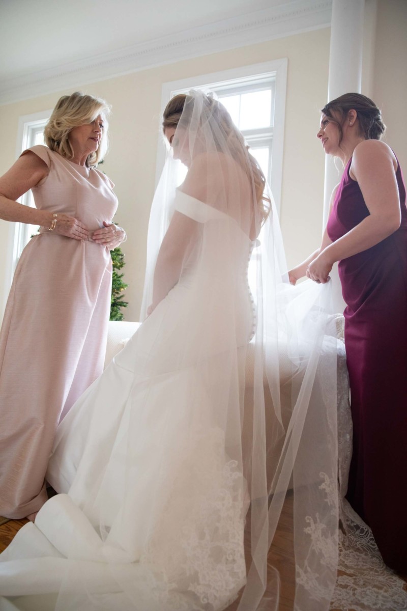 Scarlett-Michael-Wedding-Low-Res-35 - TOMMY GATZ ENTERTAINMENT Three women in a room, with one wearing a wedding dress and veil. Two women assist her, one in a light pink dress and the other in a burgundy dress.