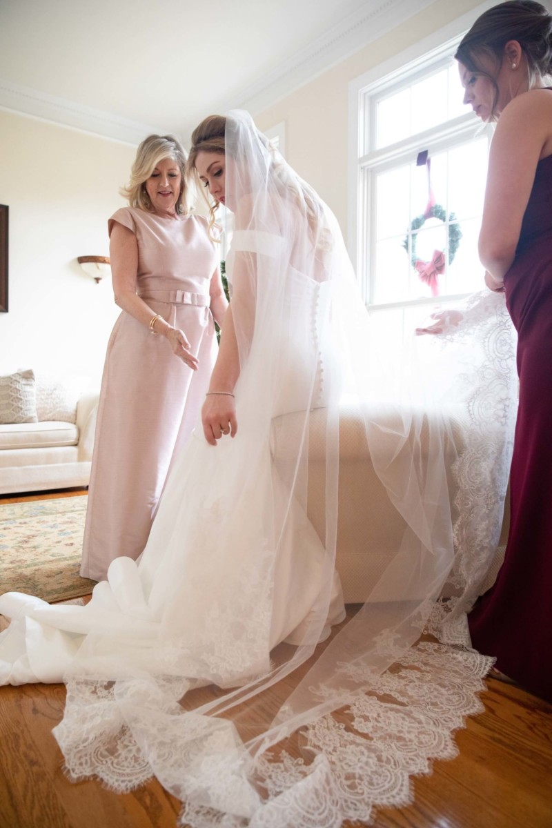 Scarlett-Michael-Wedding-Low-Res-36 - TOMMY GATZ ENTERTAINMENT Bride in white dress and veil is assisted by two women in a well-lit room with a sofa and wreath on the window.