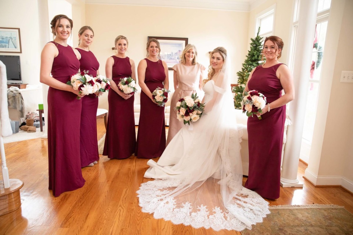 Scarlett-Michael-Wedding-Low-Res-38 - TOMMY GATZ ENTERTAINMENT Bride in white dress sits with five bridesmaids in burgundy dresses, all holding bouquets, in a living room decorated for the holidays.