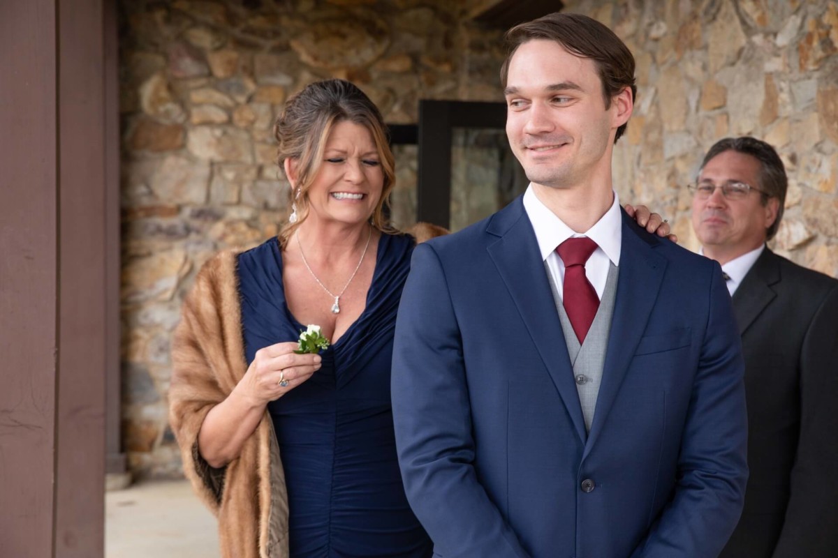 Scarlett-Michael-Wedding-Low-Res-39 - TOMMY GATZ ENTERTAINMENT A woman stands smiling behind a man in a suit while holding a boutonniere. Another man is standing in the background. They are in front of a stone wall.