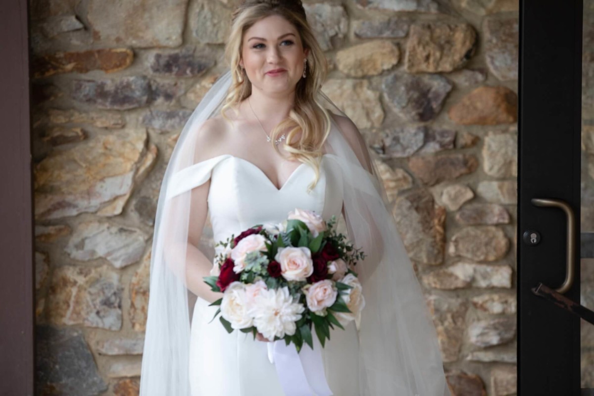 Scarlett-Michael-Wedding-Low-Res-43 - TOMMY GATZ ENTERTAINMENT A bride in a white dress holding a bouquet of pink and white flowers stands in front of a stone wall.