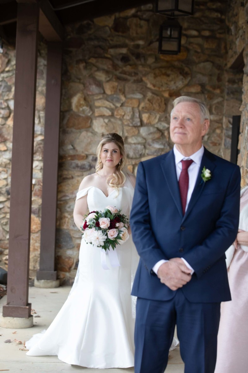 Scarlett-Michael-Wedding-Low-Res-44 - TOMMY GATZ ENTERTAINMENT A bride in a white dress holds a bouquet while standing behind an older man in a suit in front of a stone wall.