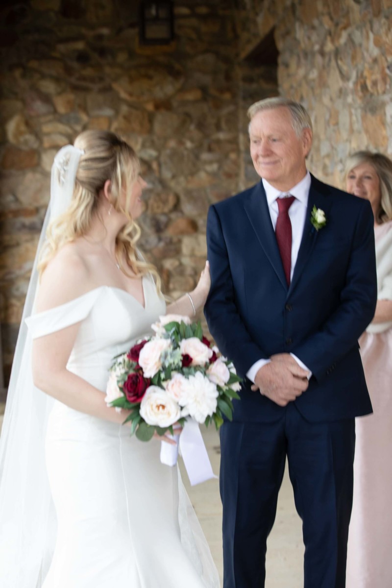 Scarlett-Michael-Wedding-Low-Res-45 - TOMMY GATZ ENTERTAINMENT A bride in a white dress holds a bouquet and stands with an older man in a suit, smiling and looking at each other. A woman in the background looks on.