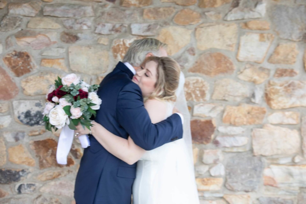 Scarlett-Michael-Wedding-Low-Res-46 - TOMMY GATZ ENTERTAINMENT Bride in white dress hugs man in suit, holding a bouquet, against a stone wall background.