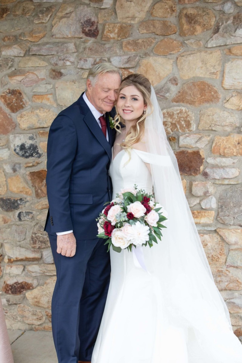 Scarlett-Michael-Wedding-Low-Res-47 - TOMMY GATZ ENTERTAINMENT A bride in a wedding dress holds a bouquet while standing beside a man in a suit. They are in front of a stone wall.