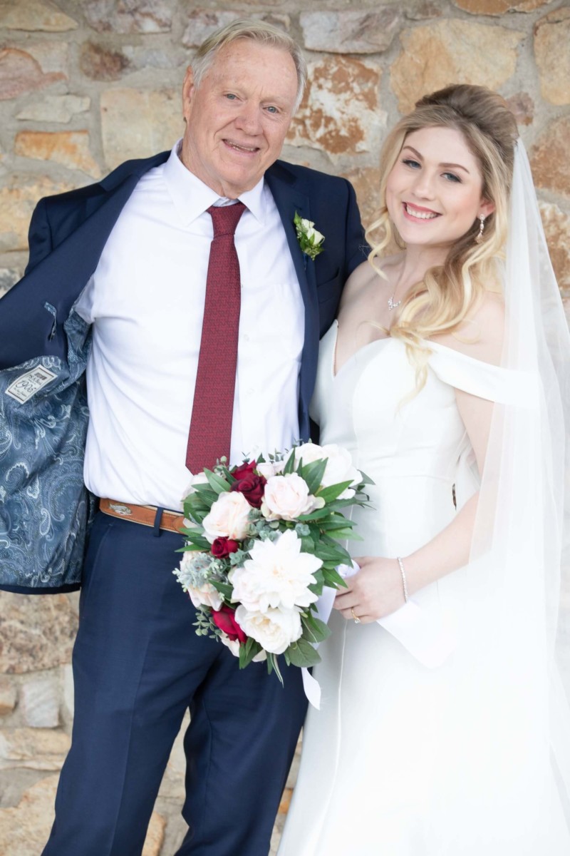 Scarlett-Michael-Wedding-Low-Res-48 - TOMMY GATZ ENTERTAINMENT A man in a suit and a woman in a wedding dress smile while standing together in front of a stone wall. She holds a bouquet of flowers.