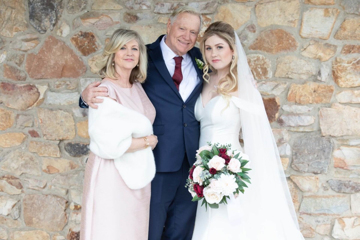Scarlett-Michael-Wedding-Low-Res-49 - TOMMY GATZ ENTERTAINMENT A bride in a white dress stands with two older adults, one in a suit and the other in a pink outfit, against a stone wall. The bride holds a bouquet.