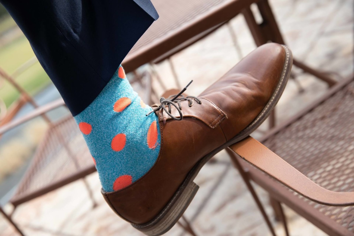 Scarlett-Michael-Wedding-Low-Res-5 - TOMMY GATZ ENTERTAINMENT Close-up of a leather shoe with blue socks featuring large orange polka dots, worn by a person on a metal chair outdoors.