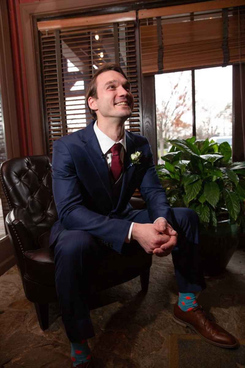 Scarlett-Michael-Wedding-Low-Res-50 - TOMMY GATZ ENTERTAINMENT A man in a navy suit and colorful socks sits on a leather chair, smiling, with a plant and window in the background.